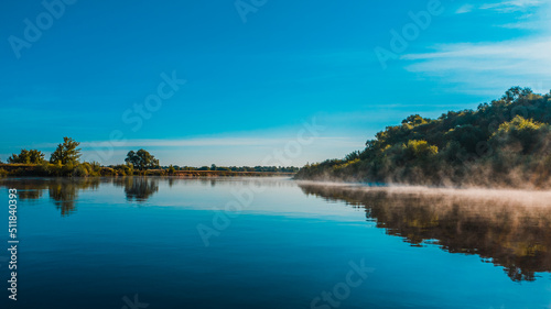 Aerial view of a beautiful summer landscape over river while dawn. Top view over river with a smooth water surface reflecting blue sky. Morning evaporation on a river while sunrise.