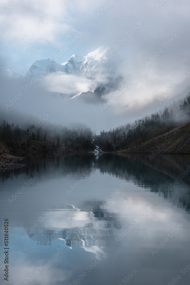 Fototapeta premium Tranquil scenery with snow castle in clouds. Mountain creek flows from forest hills into glacial lake. Snowy mountains in fog clearance, small river and coniferous trees reflected in calm alpine lake.