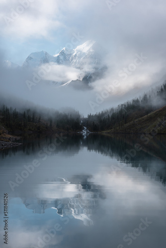 Tranquil scenery with snow castle in clouds. Mountain creek flows from forest hills into glacial lake. Snowy mountains in fog clearance, small river and coniferous trees reflected in calm alpine lake.