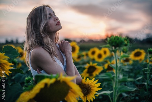 A young  slender girl with loose hair in a T-shirt stands in a field of sunflowers at sunset
