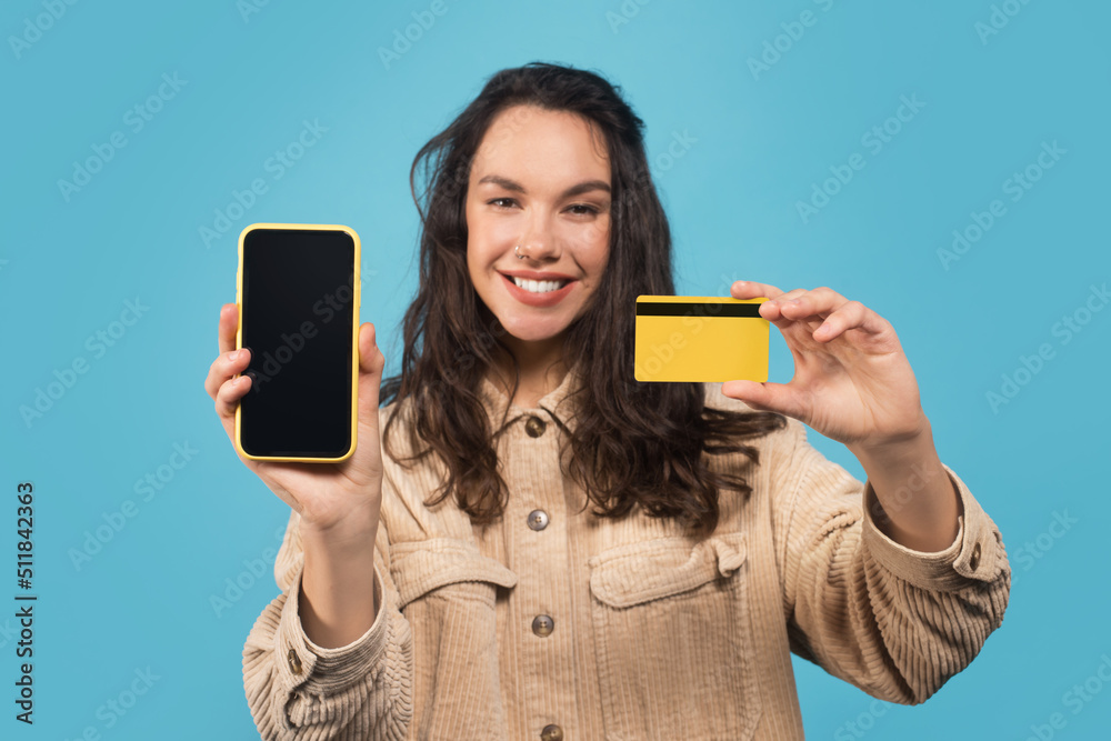 Smiling young caucasian lady student in casual showing credit card and smartphone with blank screen