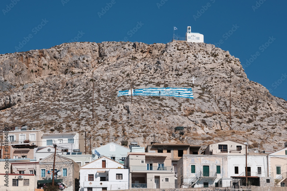 Greek national flag painted on a rock above Kalimnos town and traditional church. Kalymnos island, Greece.