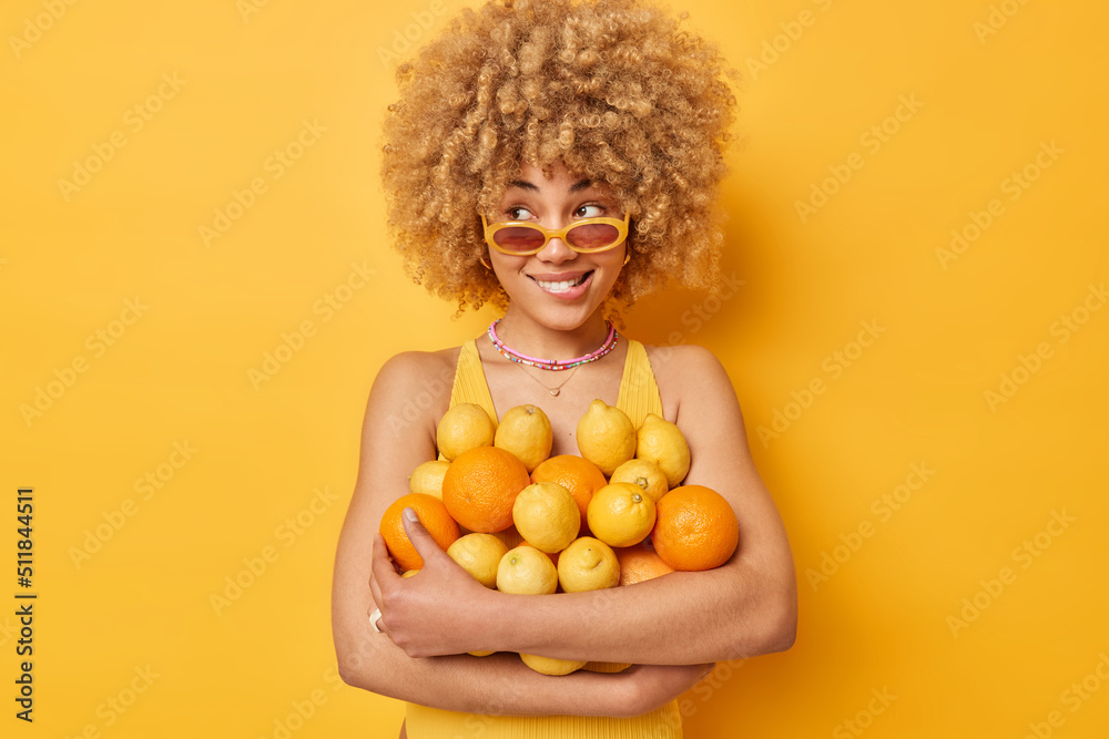 Indoor shot of pensive curly haired woman wears sunglasses embraces heap of citrus fruits eats healhy food full of vitamins poses against bright yellow background. Female model with oranges and lemons Indoor shot of pensive curly haired woman wears sunglasses embraces heap of citrus fruits eats healhy food full of vitamins poses against bright yellow background. Female model with oranges and lemons