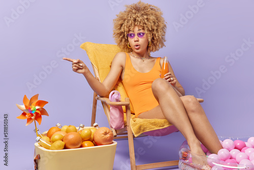 Surprised curly haired young woman points away into distance cannot believe own eyes drinks energetic cold beverage dressed in bathingsuit poses on deck chair isolated over purple background