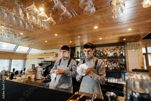 Two stylish bartenders in masks and uniforms during the pandemic, rub glasses to shine. The work of restaurants and cafes during the pandemic.. © Andrii