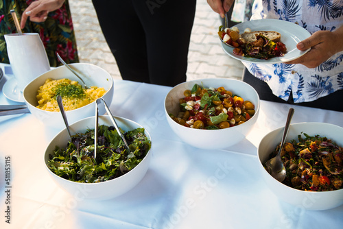 Salad bowls standing on a table for a self-serving dinner at a wedding party, or garden party. Guy with a floral shirt serving himself on a white plate. 