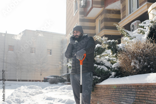A man stands with a shovel and removes snow in front of the house on a sunny and frosty day and talks on the phone. Cleaning the street from snow on a winter day.  © Andrii
