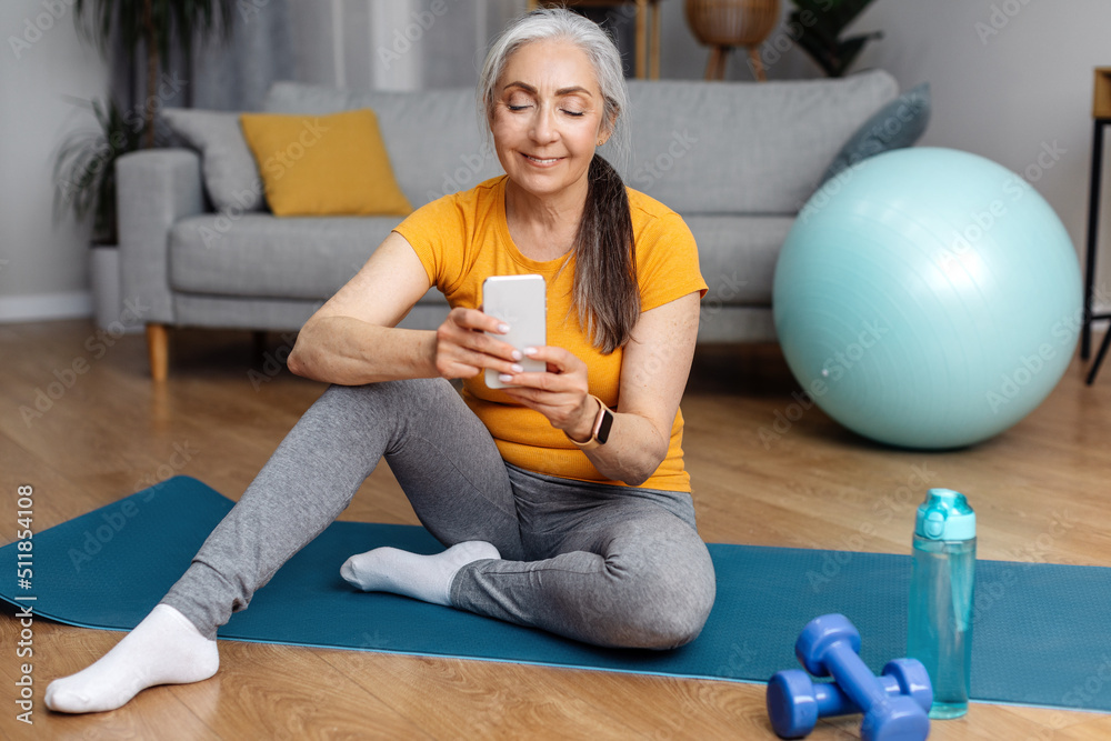 Happy athletic senior woman in sportswear sitting on yoga mat at home and  using modern cellphone Stock Photo