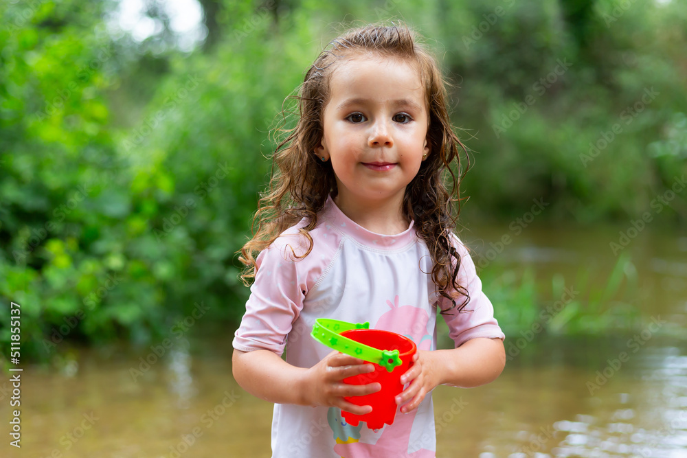 Little girl throwing water with a bucket into the river. Girl with swimsuit and t-shirt. Children's photo in summer. Little girl throwing water with a bucket into the river. Girl with swimsuit and t-shirt. Children's photo in summer.