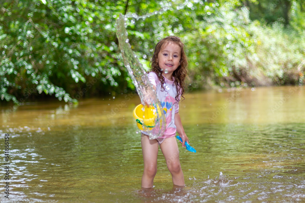 Little girl throwing water with a bucket into the river. Girl with swimsuit and tshirt