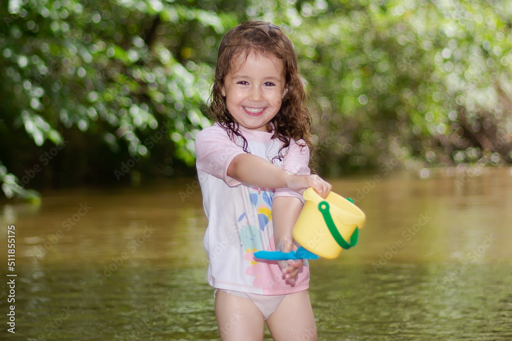 Little girl throwing water with a bucket into the river. Girl with swimsuit and tshirt