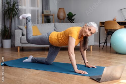 Online home fitness. Senior woman exercising to sports video on laptop in living room, looking at camera and smiling