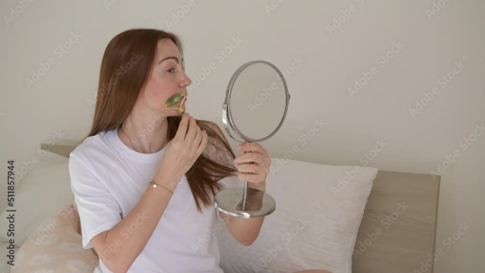 Relaxed middle age woman doing face massage with green quartz stone roller and sitting on the bed in bedroom morning. Gua sha facial massage, skin rejuvenation. Alternative beauty care, Face lift Relaxed middle age woman doing face massage with green quartz stone roller and sitting on the bed in bedroom morning. Gua sha facial massage, skin rejuvenation. Alternative beauty care, Face lift