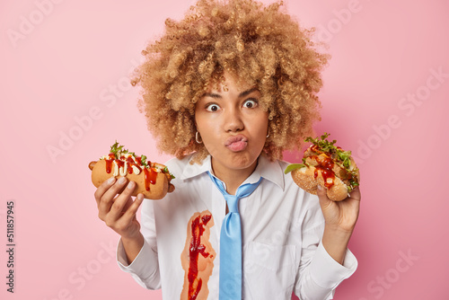 Surprised wondered woman keeps lips folded holds appetizing hot dog and hamburger prefers eating fast food dressed formally isolated over pink background. Cheat meal and unhealthy nutrition.