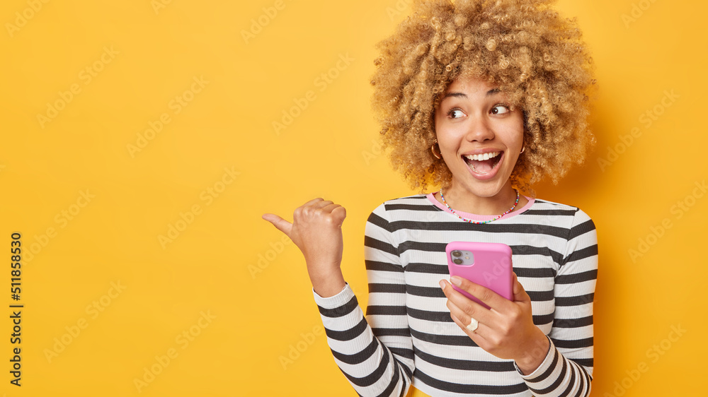 Obraz premium Horizontal shot of cheerful woman holds mobile phone sends text messages in online chat points thumb away dressed in casual striped jumper isolated over yellow background shows announcement.