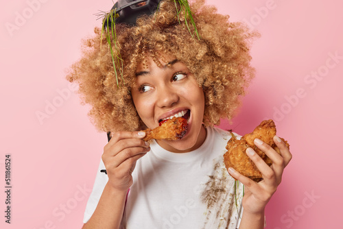 Beautiful woman feels very hungry eats chicken nuggets with ketchup wears dirty t shirt and protective helmet enjoys harmful fast food looks away isolated over pink background. Unhealthy eating