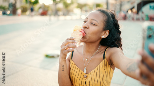 Closeup of smiling young woman with long curly hair with ice cream in her hands making video call on a mobile phone on an urban city background. Close-up of happy girl using smartphone to communicate