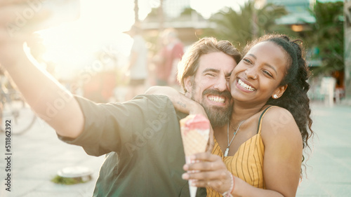 Closeup of smiling interracial couple eating ice cream and taking a selfie on urban city background. Close-up of a man and woman tasting ice cream and video chatting using a mobile phone. Backlight