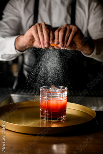 Close-up of glass with alcoholic beverage and male hands carefully sprinkle by citrus zest juice on it