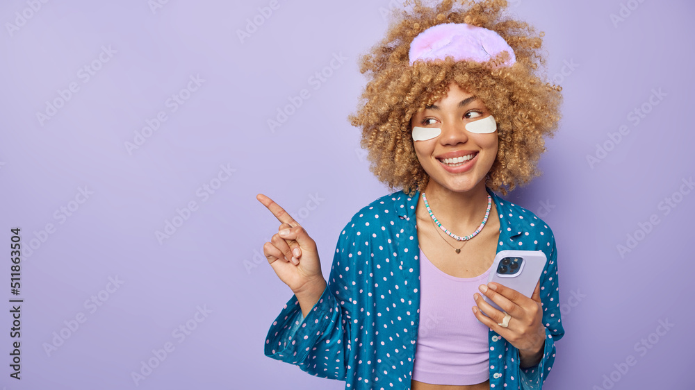 Horizontal shot of cheerful curly haired young woman dressed in nightwear applies beauty patches to reduce puffiness holds mobile phone point left on empty space isolated over purple background Horizontal shot of cheerful curly haired young woman dressed in nightwear applies beauty patches to reduce puffiness holds mobile phone point left on empty space isolated over purple background