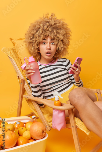 Shocked curly woman dressed casually holds bottle of water and cellphone stares impressed poses on comfortable deck chair spends summer vacation on beach uses portable freezer for cooling fruits