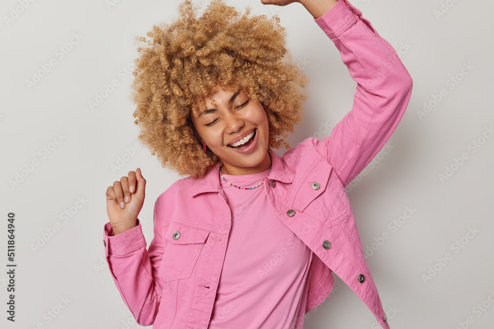 Waist up shot of cheerful European woman with blonde curly hair dances carefree shakes arms has upbeat mood dressed in pink jacket smiles broadly isolated over grey background. Winner dance. Waist up shot of cheerful European woman with blonde curly hair dances carefree shakes arms has upbeat mood dressed in pink jacket smiles broadly isolated over grey background. Winner dance.