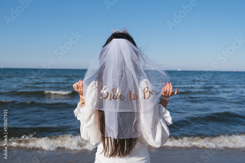 Fototapeta Naklejka Na Ścianę i Meble -  Bride standing from the back to camera with veil infront of sea. Bride in white dress on the beach. Bride to be. Bachleorette party on the beach