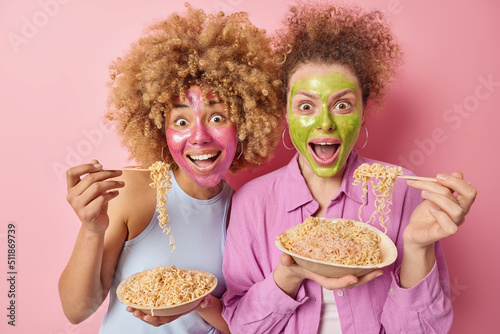 Two surprised young women with curly hair apply beauty maskon face for skin moisturising eat delicious pasta undergo facial treatments dressed in casual clothes isolated over pink background