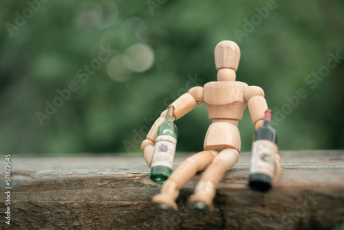 Wooden mannequin doll sitting on a bench with two bottles of vine, like a drunk person. Photo on a natural background with bokeh, horizontal photo
