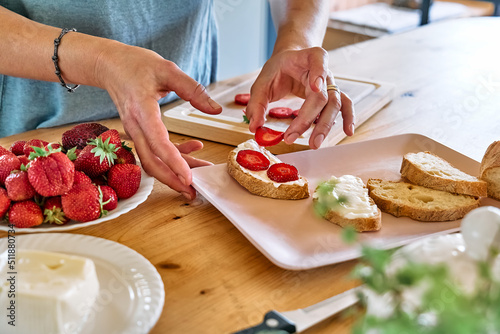 Woman making summer strawberry sandwich. Female hands putting slices of ripe red strawberry on toast with spread stracchino cheese. Healthy eating, fruit dieting brunch.