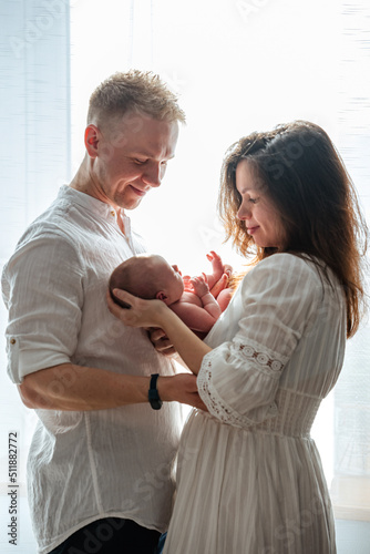 Dad and mom hug a newborn baby at home, young parents happily look at their child. contour light from the window