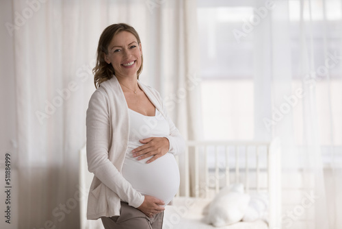 Expectant mother portrait concept. Beautiful young 30s pregnant woman in casual comfort clothes posing standing in modern nursery touch big belly in late pregnancy smiling looking at camera feel happy