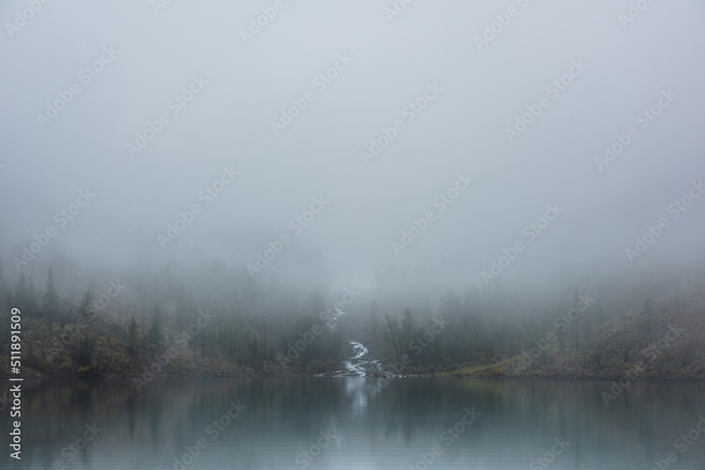 Mountain creek flows from forest hills into glacial lake in mysterious fog. Small river and coniferous trees reflected in calm alpine lake in misty morning. Tranquil landscape in fading autumn colors.