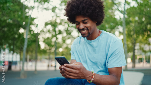 Closeup portrait of young African American man in light blue t-shirt sitting on city park bench and using his smartphone. Man looks at photos, videos in his mobile phone. lifestyle concept.