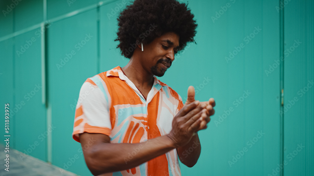 Close-up of cheerful young African American man wearing shirt listening to music in wireless headphones and dancing on light blue wall background. Lifestyle concept. - obrazy, fototapety, plakaty 
