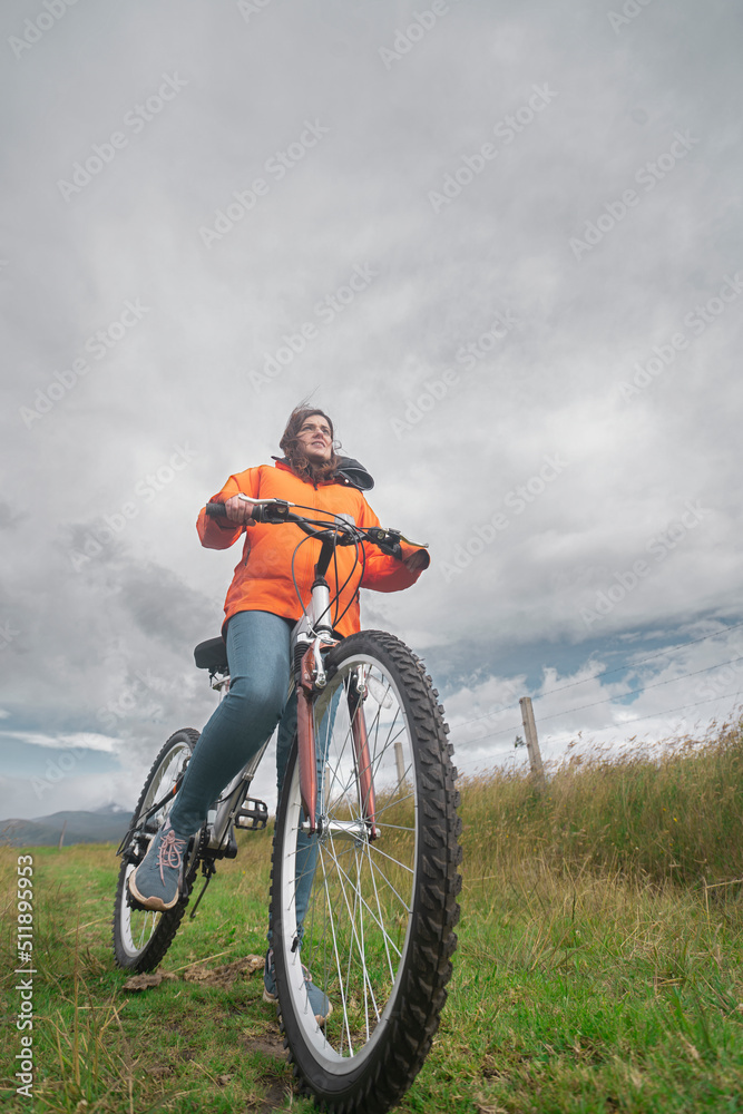 Young Hispanic woman wearing orange jacket and blue pants riding her bike through rural countryside