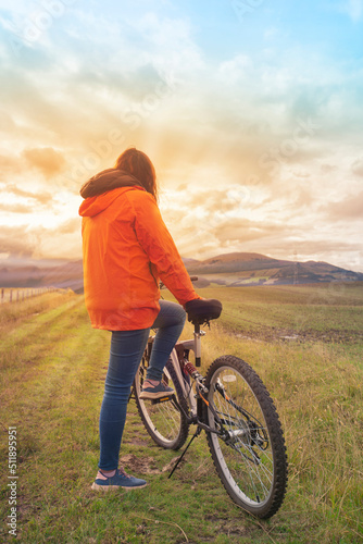 Young Hispanic woman wearing an orange jacket and blue pants resting leaning on her bicycle in the middle of a rural field during a sunset