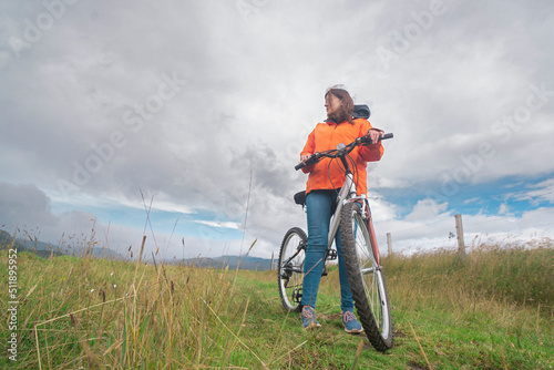 Fototapeta Naklejka Na Ścianę i Meble -  Young Hispanic woman wearing orange jacket and blue pants resting on her bicycle in the middle of a rural field during a cloudy day