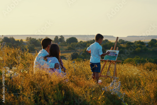 Family wearing summer outfits relaxing at sunset on hill. Parents sitting in tall grass and watching little son drawing picture on canvas. Creative hobby. Concept of arts