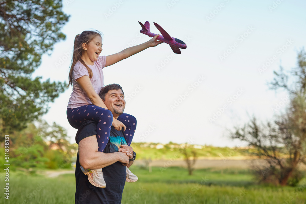 Fototapeta premium Cute girl riding on father's shoulder playing with toy airplane