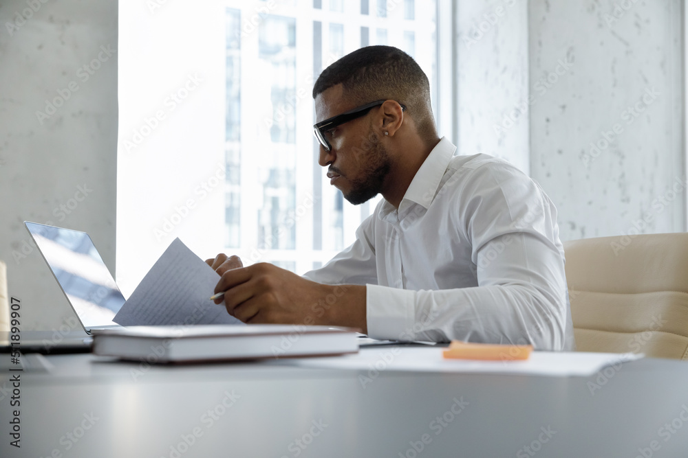 Young serious African businessman, accountant or lawyer do paperwork, working on documents uses laptop, checks terms of agreement, prepare contract looking focused seated at modern office workplace Young serious African businessman, accountant or lawyer do paperwork, working on documents uses laptop, checks terms of agreement, prepare contract looking focused seated at modern office workplace