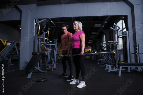 Full length shot of an elderly woman lifting barbell, working out with personal trainer at the gym
