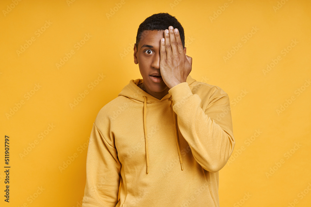 Shocked mixed race man covering half face with hand while standing against yellow background Shocked mixed race man covering half face with hand while standing against yellow background