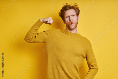 Konstfotografi Playful redhead man showing his bicep while standing against yellow background