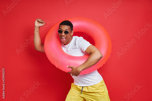 Happy mixed race man carrying inflatable ring while standing against red background