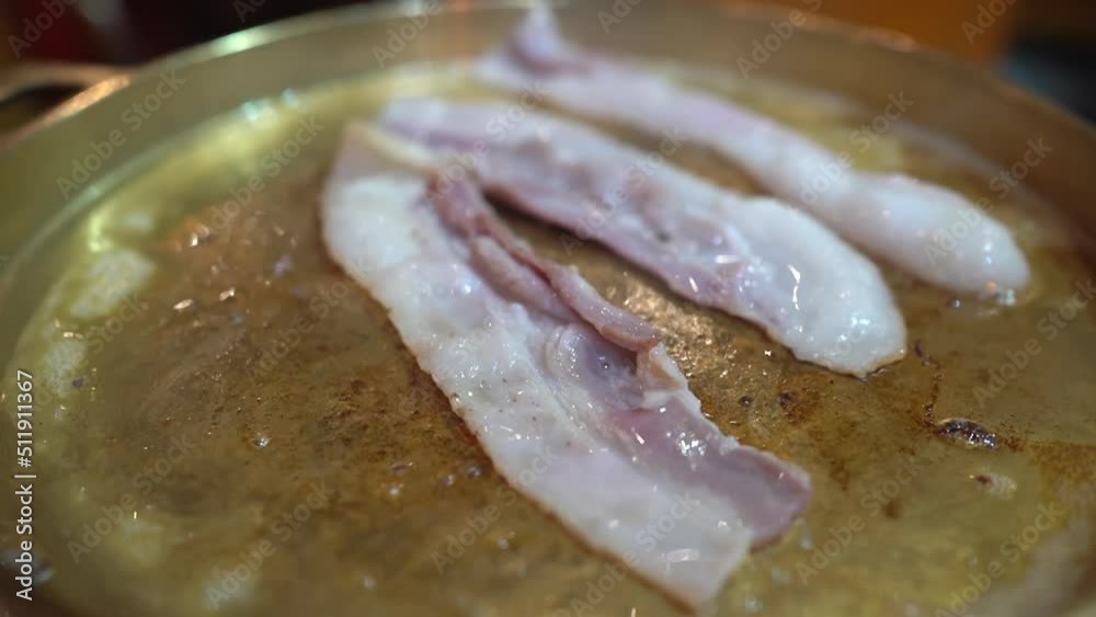 Close-up of Moo Ka Ta, Thai BBQ buffet with three pieces of sliced bacon are grilled on the BBQ grill pan with little boiling oil from the bacon in BBQ restaurant. Focus on the first piece of bacon.