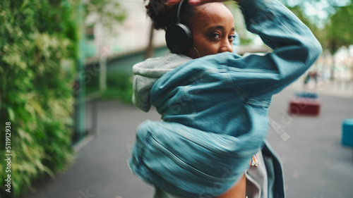 Closeup, smiling African girl wearing denim jacket, in crop top with national pattern walks in city park listening to music on headphones and dancing on green trees background.