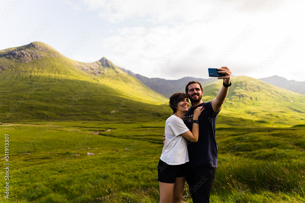 Couple taking a selfie with a beautiful landscape full of mountains on a summer day in the highlands, Scotland.