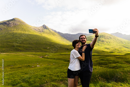 Couple taking a selfie with a beautiful landscape full of mountains on a summer day in the highlands, Scotland.