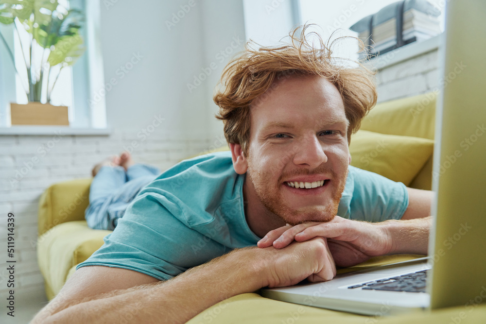 Cheerful redhead man using laptop and smiling while lying on the couch at home Cheerful redhead man using laptop and smiling while lying on the couch at home